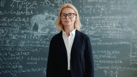 Portrait of mature adult woman wearing suit standing alone in class, smiling and looking at camera, blackboard with formulas is visible in background.