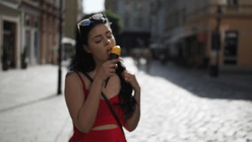 Young beauty woman in red dress is smiling and eating ice cream during walking in the street of historical part of Bratislava city in sunny summer afternoon. Half speed. 
Close up.