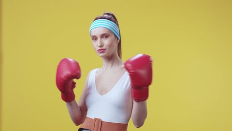 Strong and beautiful sports woman kickboxing with red gloves forward doing some boxing exercises smiling joyful at camera. Yellow wall. Healthy life, activities and sports.