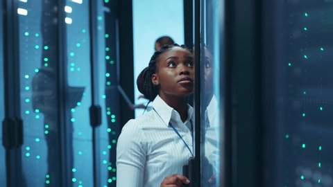 African american woman engineer working in business team of data center inspecting open server racks and typing data on tablet. People and technology.