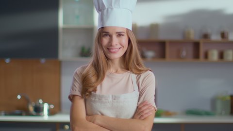 Portrait of smiling chef woman standing on kitchen. Lady in apron smiling at camera indoor in slow motion. Woman in chef hat crossing hands on luxury kitchen