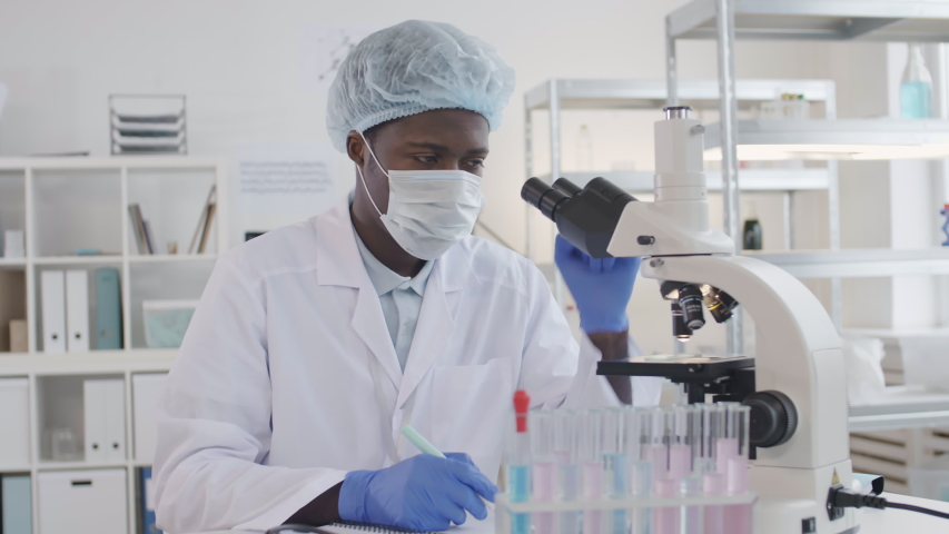 Medium shot of African scientist wearing medical overall, protective hat, mask and gloves making research using microscope while his colleague working on background | Shutterstock HD Video #1046833789