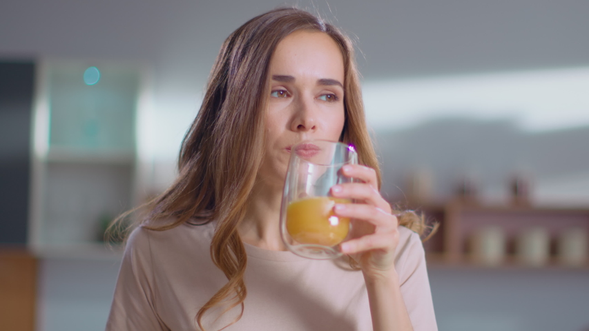 Close up of woman pouring orange juice into the glass from jar on kitchen in morning. Girl holding glass of fruit juice at home. Smiling lady drinking orange juice on modern kitchen in slow motion