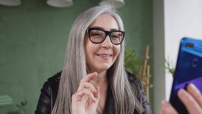 Beautiful satisfied retired stylish lady in eyeglasses with long gray hair having video chat and dreamly looking aside after she finished the conversation