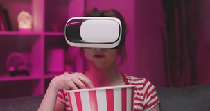 Young woman wear VR headset and watch movie with popcorn at night. American woman sitting on the sofa in the VR glasses and watching something while eating popcorn. Indoor