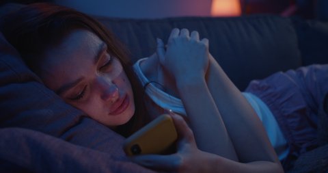 Close up of lonely young woman lying on sofa and using smartphone at night. Depressed millennial female starring at mobile phone screen and thinking. Concept of painful brokeup.