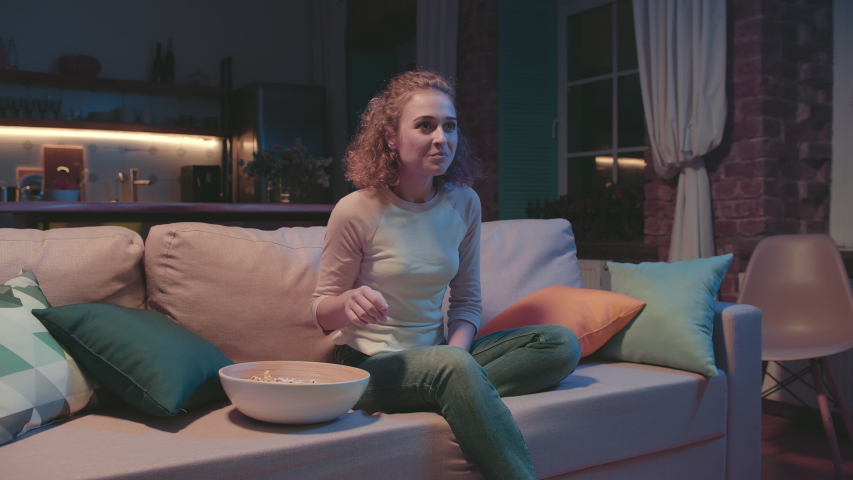 Portrait of young cheerful woman sitting on sofa, eating popcorn while watching favorite romantic tv show and clapping her hands with excitement and joy. Royalty-Free Stock Footage #1046848777