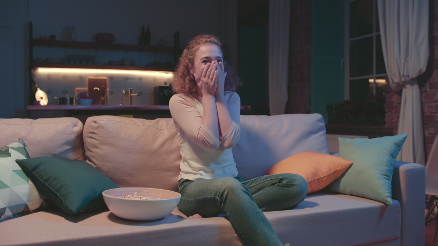 Portrait of young cheerful woman sitting on sofa, eating popcorn while watching favorite romantic tv show and clapping her hands with excitement and joy.