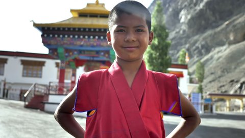 Close up shot of a young Tibetan Buddhist monk in a red robe looking into a camera with a smile while standing next to a monastery situated in Himalaya mountain.