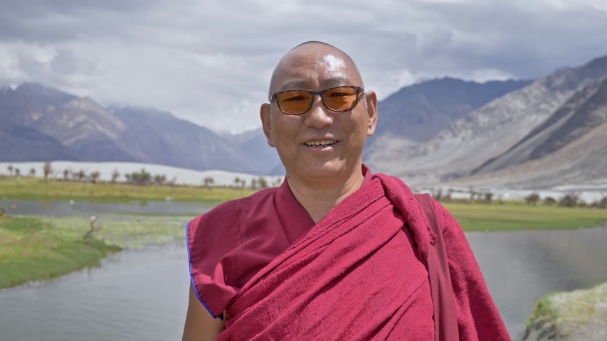 A happy Buddhist monk in red robe wearing eyeglasses smiling looking at the camera. A buddhist monk standing beside a river and lush green pasture situated in Himalaya mountains in Ladakh, India | Shutterstock HD Video #1046849617