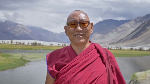 A happy Buddhist monk in red robe wearing eyeglasses smiling looking at the camera. A buddhist monk standing beside a river and lush green pasture situated in Himalaya mountains in Ladakh, India