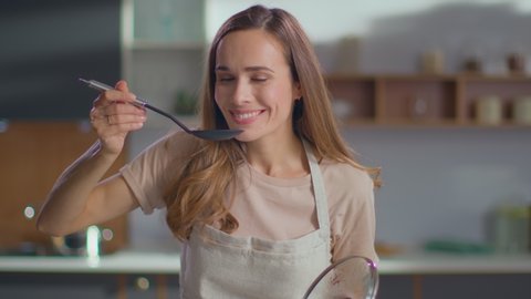 Pretty woman cooking soup on stove at kitchen. Close up of housewife preparing dinner at home. Young woman tasting soup on spoon at modern kitchen in slow motion