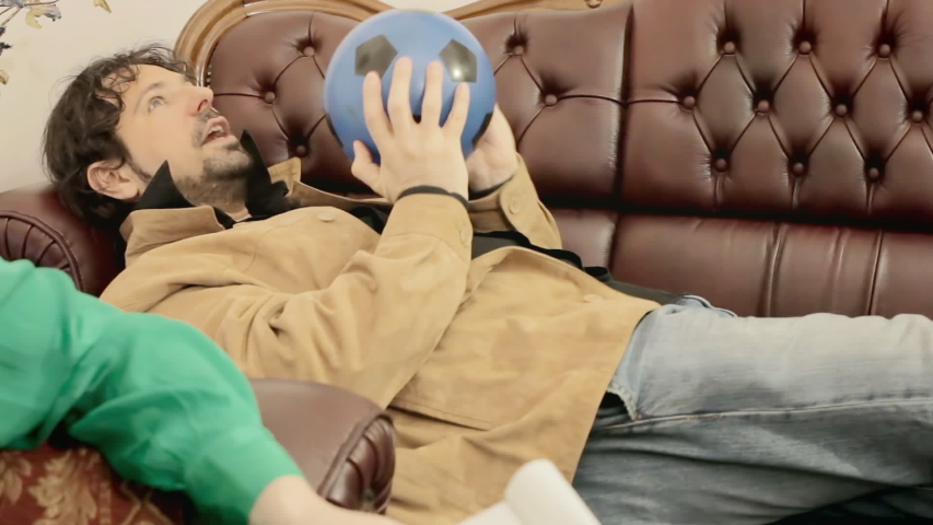 A man talking to a psychotherapist while lying on a couch and playing with a blue soccer ball. Odd fixation. Medium shot.
