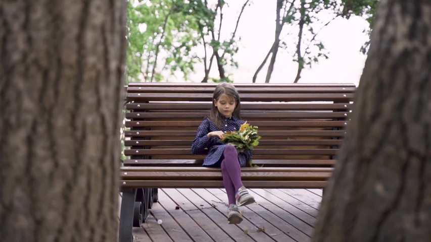 Little beautiful girl in a blue dress with leaves in her hands laughs on a bench in the park