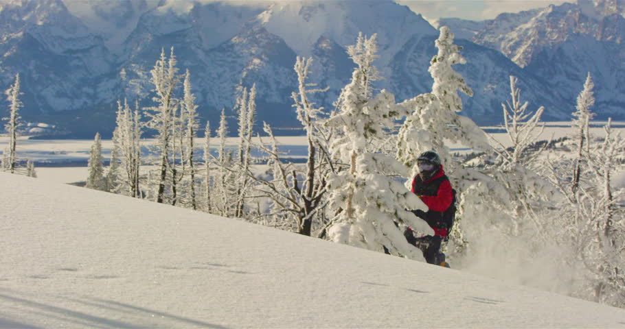 Man snowmbiles up hill and makes powder turn with Grand Teton in background.
