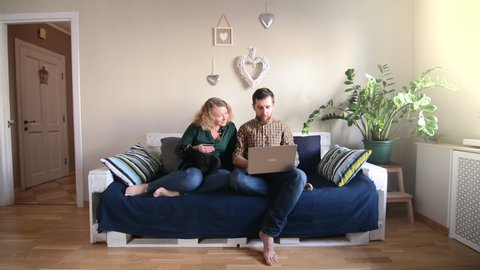 Young Caucasian couple doing shoppings online on the couch at home with laptop, looking at each other and smiling