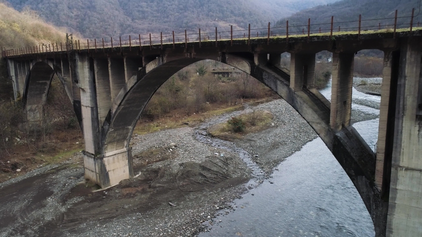 Flying under rusty bridge crossing the narrow river lwith stony bottom surrounded by mountains and forest. Shot. Aerial of rural landscape in late autumn.