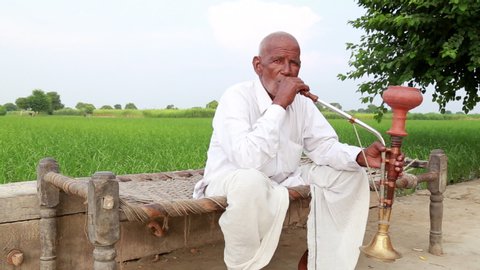 Sonepat, India - August 9, 2014:Old Men Sitting on Coat and Enjoying Hookah in the Green Field. He is Wearing Indian Traditional Clothes Dhoti and Kurta.