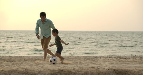 Asian Father and son playing together with ball in football on the beach under sunset background. Slow motion. Happy family concept. 