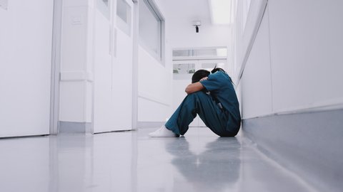 Stressed and overworked female doctor wearing scrubs sitting on floor in hospital corridor - shot in slow motion