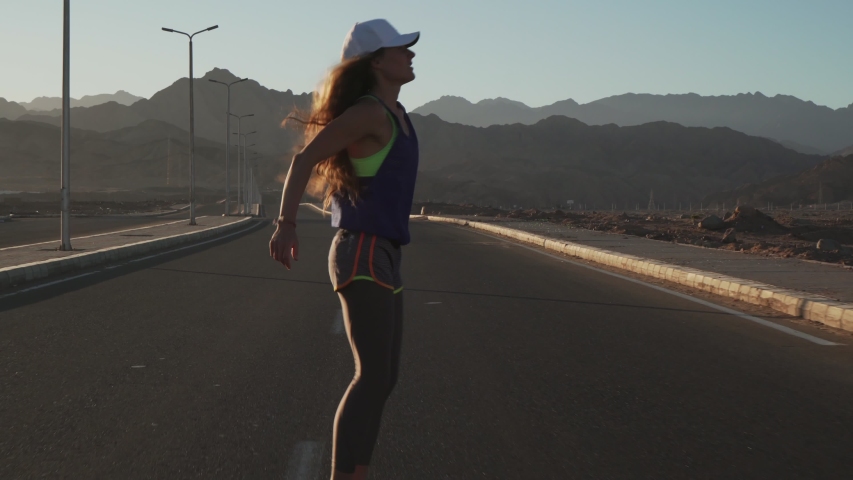 Young motivated girl doing jumping exercise during jogging at highway at sunset