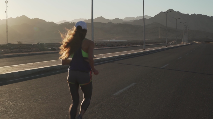 Back view of young slim girl jogging at highway in mountains at sunset