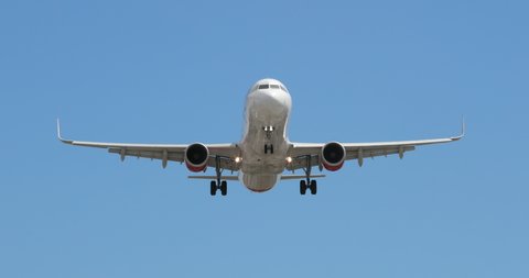 Frontal shot of modern white airplane landing, with landing gears seen released. Clear blue sky is all around the plane. Airliner smoothly descends approaching the camera and flies right above it. 4K