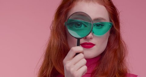 Funny colorful Girl looking throw Magnifier to the Camera in Image Sunglasses. Trendy red-haired Woman having fun. Colorful Portrait. Beautiful Girl. Funny Frame. Amazing Eyes. Closeup Face.