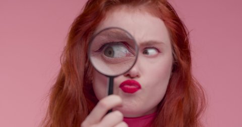 Funny red-haired Woman looking throw Loupe comically and shocked, chewing gum. Attractive young Woman with bright pink Makeup showing funny Emotions in Frame. Looking funny.Having Fun.Shocked Woman.