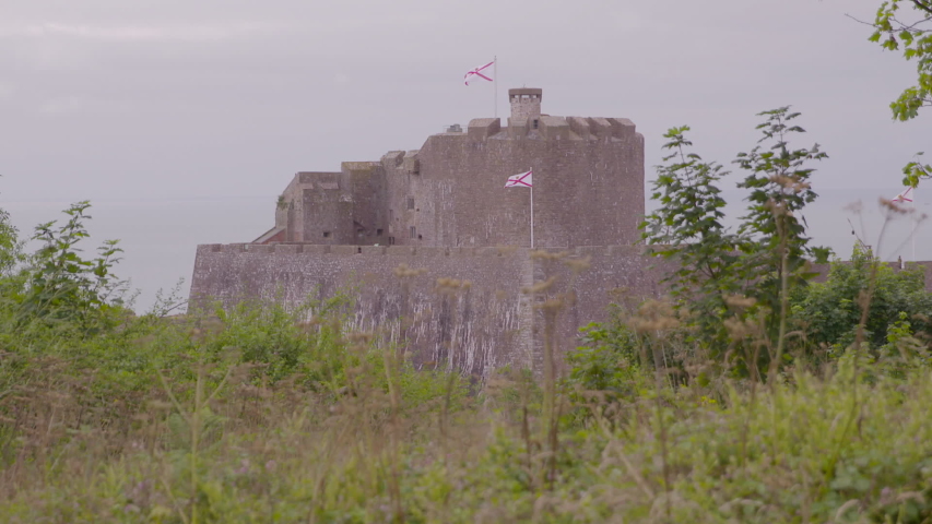 Wide view of Mont Orgueil Castle, Saint Martin, Jersey, showing flag of Jersey flying, with trees and bushes in foreground