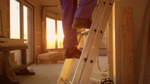 SLOW MOTION, CLOSE UP, LENS FLARE, DOF: Builder in blue overalls climbs up a aluminium ladder at sunset. Unrecognizable construction site worker climbs up an extendable ladder on a sunny evening.