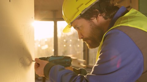 SLOW MOTION, LENS FLARE, CLOSE UP: Young contractor working late uses a power drill to mount a wall panel. Bearded worker changes bolts fastening a gypsum wallboard. Builder working on a sunny evening