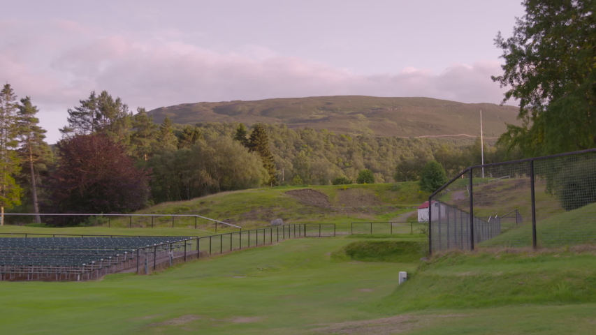 Medium low angle still shot of a fenced Scottish sports playground, outer pine trees, and horizon mountain landscape against the cloudy sky, Braemar, Scotland, UK