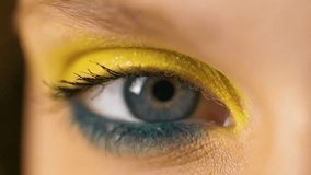 Close up of beautiful Woman’s eye looking at Camera objective, having long Eyelashes. Woman with colorful makeup. Yellow and blue eyeshadow. Attractive Appearance. Blue eye of female model.
