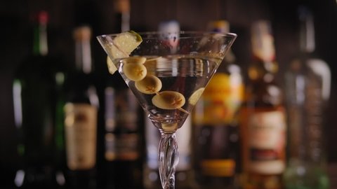 Close-up of a cool cocktail with vermouth vodka or gin with green olives and lime stands on a rotating surface against the background of a bar with bottles. Slow motion.