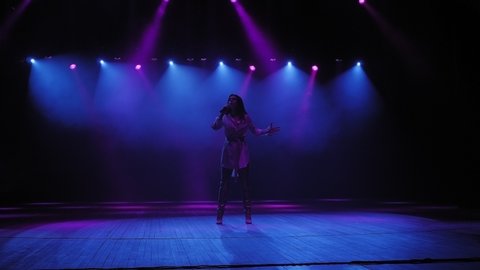 Silhouette of a beautiful girl singer with microphone in the dark. A gorgeous girl singer sings on stage in the dark under the light of blue and purple floodlights. Slow motion.