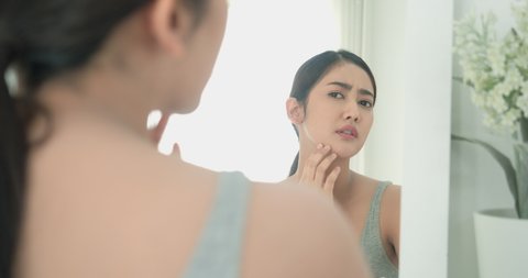 Happy confident Asian beautiful young woman looking face at mirror and worry facial skin problems in the morning at home. Beauty girl has problems face skin.