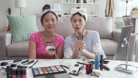 Two beauty blogger cute asian girls presenting beauty cosmetic products and broadcasting live video to social network. female vloggers face camera talking while holding makeup palette and brushes