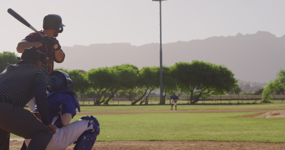 Side view of a Caucasian male baseball player during a baseball game, hitting a ball with a baseball bat and running on a sunny day, with other players in the foreground, in slow motion