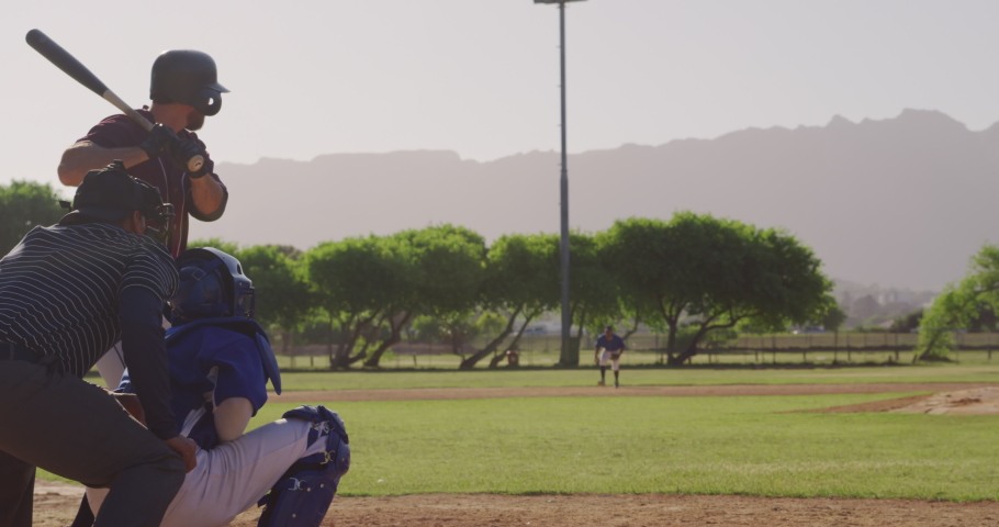 Side view of a Caucasian male baseball player during a baseball game, hitting a ball with a baseball bat and running on a sunny day, with other players in the foreground, in slow motion Royalty-Free Stock Footage #1046954011