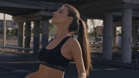 A concentrated calm young woman in sports bra is stretching her back before training outdoors near the bridge in the city