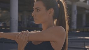 A lovely calm young woman in sports bra is stretching her arms before training then looking to the camera outdoors near the bridge in the city