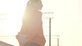 A slim young woman wearing an orange hoodie is stretching her leg outdoors on the bridge in the sunny morning