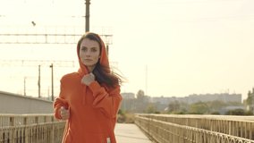 A slim focused young woman in an orange hoodie is run outdoors on the bridge in the early morning