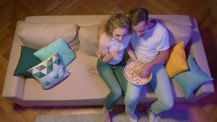 Top view of young lovely couple with popcorn watching comedy show on tv and laughing enjoying free evening together at home. Young man and woman enjoying tv series on sofa