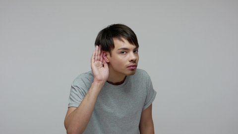 Young guy with hearing problems holding hand near his ear and listening around carefully, being focused attentive to sounds, trying to hear conversation. indoor studio shot isolated on gray background
