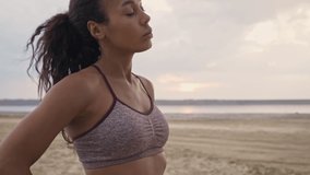 A calm young african american woman in sportswear is warming up on the beach in the morning