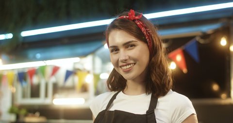 Portrait of attractive Caucasian joyful young girl in apron smiling happily to camera outdoor with small food truck on background. Close up of pretty female bartender. Woman vendor at fair in park.