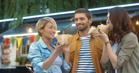 Caucasian handsome guy and two pretty girls eating hot dogs outdoor at food court in park. Women feeding man with fast food at outside bar. Happy cheerful friends.