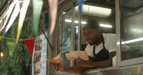 African American joyful young man barman looking out window of festive food truck with two hot dogs in hands. Male waiter smiling while giving order. Handsome cheerful vendor of small cafe.